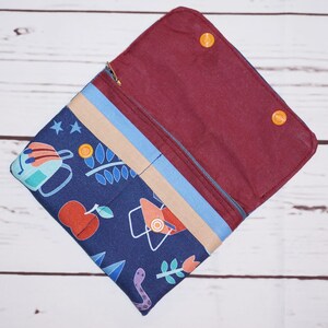 May include: Open, navy blue fabric wallet with a camping-themed pattern. The wallet features a zipper closure, a tan and light blue stripe, and a burgundy interior. Orange snap closures secure the wallet.