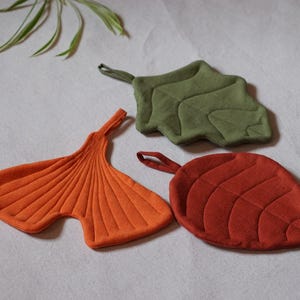 May include: Three leaf-shaped pot holders in shades of orange, green, and red. Each has a loop for hanging. The orange one is a ginkgo leaf, the green one is a maple leaf, and the red one is a broad leaf.