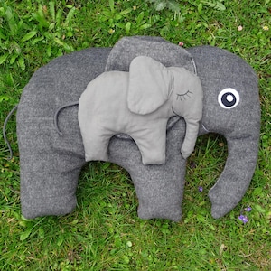 Op de afbeelding: Een grijs olifantvormig kussen met een kleiner grijs olifantenkussen erop. Het grote kussen heeft een wit oog en een slurf. De kleinere olifant heeft gesloten ogen. De kussens liggen op groen gras.