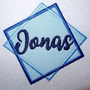 May include: A square-shaped embroidered patch featuring the name Jonas in dark blue cursive script. The patch has a light blue fabric base with a dark blue stitched border, layered over a second identical light blue square patch.