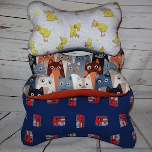 Könnte beinhalten: Drei knochenförmige Kissen übereinander gestapelt. Das obere Kissen ist hellgrau mit gelben Entenküken und floralen Akzenten. Das mittlere Kissen hat ein Muster aus verschiedenen Katzenköpfen. Das untere Kissen ist marineblau mit einem rot-weißen Muster.