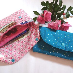 May include: Two fabric wallets. One is open, showing a pink floral pattern, a zipper, and contents like a phone and cards. The other is closed, teal with a triangle pattern and a button closure. Pink roses are in the background.