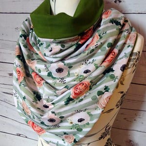 Peut inclure: Un foulard floral avec un col vert. Le foulard présente un motif de roses roses et d'anémones blanches sur un fond à rayures vert clair et blanc. Le foulard est drapé sur un mannequin.