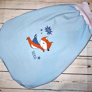 Könnte beinhalten: Hellblauer Babyschlafsack mit einem Comic-Fuchs mit Umhang. Der Fuchs ist orange und weiß mit einem blauen Umhang. Die Worte "SUPER" und "HERO" sind auf dem Schlafsack aufgedruckt. Der Schlafsack hat eine weiße Einfassung.