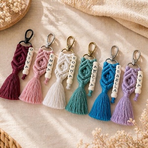May include: A collection of handmade macrame keychains in various colors, including burgundy, pink, white, teal, blue, and purple. Each keychain features a tassel and a wooden bead with a name on it. The keychains are attached to metal clasps.