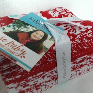 May include: A stack of red and white patterned fabric squares, tied with a white ribbon. The fabric features a floral design. Attached tags read "So Ruby" and "Riley Blake Designs".
