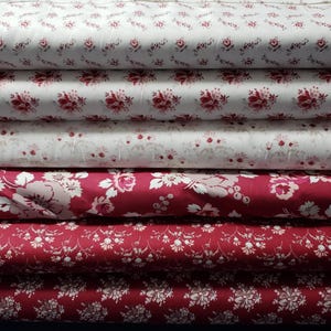 May include: A stack of fabric bolts featuring floral patterns in shades of red and white. The designs vary from small, delicate sprigs to larger, more prominent blooms. The fabric appears to be cotton or a similar material, suitable for quilting or crafting projects.