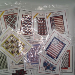 May include: A collection of Fabric Cafe quilt patterns in plastic packaging. The patterns feature various designs, including checkerboard, stripes, and patriotic themes. The patterns are labeled with names like "Freedom" and "Courage".