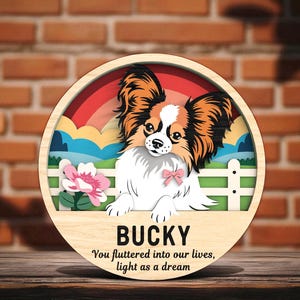 May include: A wooden, circular sign featuring a Papillon dog illustration with a rainbow and floral background. The sign includes the name "BUCKY" and the text "You fluttered into our lives, light as a dream."