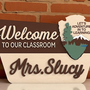May include: Wooden classroom sign with the words "Welcome to our Classroom" and "Mrs. Slucy" in a script font. The sign has a brown and beige color scheme with a decorative arrowhead shape featuring a mountain and tree scene.