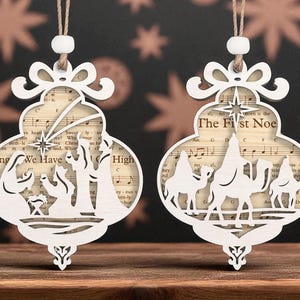 Puede incluir: Dos adornos navideños de madera blanca con un diseño recortado. Uno presenta una escena de la natividad con el texto "Angels We Have Heard on High". El otro muestra a los tres reyes magos con el texto "The First Noe."