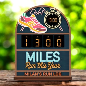 Op de afbeelding: Houten hardlooplog met een roze en gele hardloopschoenafbeelding, een stopwatch met 13:00 en een digitaal display met "1300 MILES Run this Year". De log is gelabeld "MILAN'S RUN LOG".