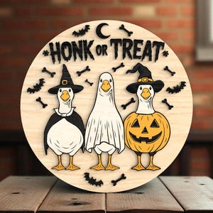 Puede incluir: Decoraci&oacute;n redonda de Halloween de madera con tres gansos disfrazados. Un ganso es una bruja, otro es un fantasma y otro es una calabaza. Las palabras "HONK OR TREAT" est&aacute;n en la parte superior, con murci&eacute;lagos y huesos alrededor.