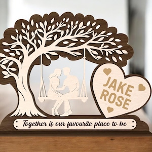 Peut inclure: Décoration en bois avec un arbre et un couple sur une balançoire. Un cœur avec les noms "Jake Rose" et la phrase "Together is our favourite place to be" complète le design.
