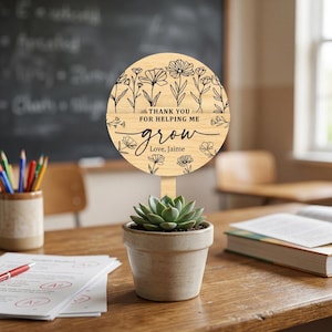 May include: A wooden plant stake with the text "THANK YOU FOR HELPING ME grow Love, Jaime" and floral illustrations. The stake is placed in a small, beige ceramic pot with a succulent plant. The scene is set on a wooden desk in a classroom.