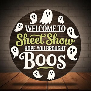 Puede incluir: Cartel de madera redondo con el texto "WELCOME TO Sheet Show HOPE YOU BROUGHT BOOS" en letras blancas y verdes. Figuras de fantasmas blancos salpican el cartel, que se encuentra sobre un fondo de madera oscura.