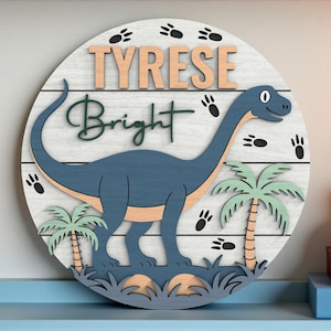 May include: A circular wooden sign with a blue dinosaur, tan belly, and green palm trees. The sign displays the name "TYRESE Bright" in wooden letters. The background is white with black paw prints.
