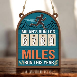 Op de afbeelding: Houten bord met de tekst "MILAN'S RUN LOG" en "6789 MILES RUN THIS YEAR." Het bord toont een silhouet van een hardloper, bergen en voetafdrukken. Het bord is bruin, blauwgroen en oranje.
