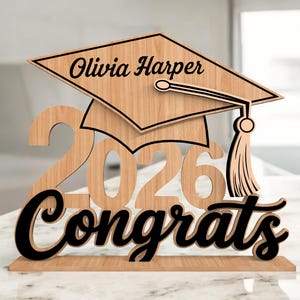 Puede incluir: Decoración de graduación de madera con el nombre "Olivia Harper" en un gorro de graduación, el año "2026" y la palabra "Felicidades" en cursiva. El diseño es en tonos marrones y negros.