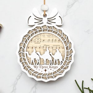 May include: A white, laser-cut Christmas ornament with a bow-shaped top. The ornament features a circular design with a musical score background and silhouettes of three camels. The text "We Three Kings" is visible.
