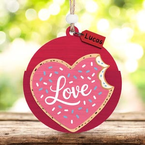 Puede incluir: Adorno de madera rojo con un diseño de donut en forma de corazón. El donut rosa tiene chispas azules y blancas y la palabra "Love" en letras blancas. Una pequeña etiqueta con el nombre "Lucas" cuelga del adorno.