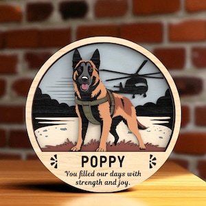 Puede incluir: Letrero redondo de madera con un perro pastor belga malinois y un helicóptero de fondo. El perro lleva un arnés. El letrero tiene el nombre "POPPY" y el texto "You filled our days with strength and joy."