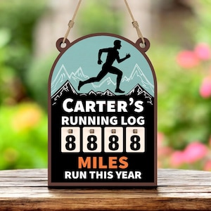 Op de afbeelding: Houten bord met een hardloopfiguur, bergen en de tekst "Carter's Running Log". Het bord toont "8888 Miles Run This Year". Het heeft een bruine rand en is opgehangen met natuurlijk touw.