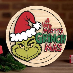 May include: A circular wooden sign with the Grinch wearing a red Santa hat and the text "A Very Merry Grinch Mas" in red and green. The Grinch's face is green with yellow eyes. The sign has a black border.