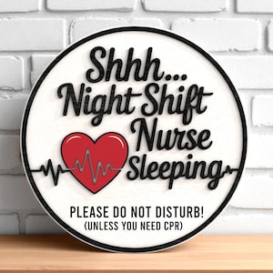 以下が含まれることがあります： 「Shhh... Night Shift Nurse Sleeping」と黒い文字で書かれた丸い白い木製の看板。心拍ラインのある赤いハートが中央にあります。下部のテキストは「PLEASE DO NOT DISTURB! (UNLESS YOU NEED CPR)」と書かれています。