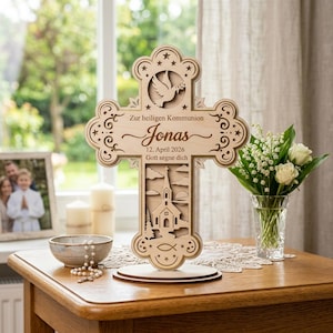 May include: A wooden cross with decorative cutouts, including a dove, stars, and a church scene. The cross has the name "Jonas" and the date "12. April 2026" engraved on it. It stands on a wooden base.