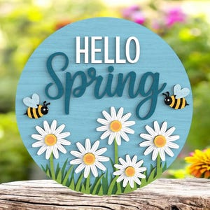 May include: A round, light blue wooden sign with the words "HELLO SPRING" in white and teal. The sign features daisies, grass, and two cartoon bees. The sign is placed on a wooden surface.