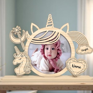 May include: Wooden photo frame with a unicorn design. The frame has a cutout for a photo and includes spaces for a name and height. The frame is decorated with a rainbow, a crescent moon, a star, and a unicorn.