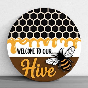 May include: Round wooden sign with a honeycomb pattern, dripping honey, and a bee illustration. The sign reads "Welcome to our Hive" in black and yellow lettering. The sign is a decorative item.