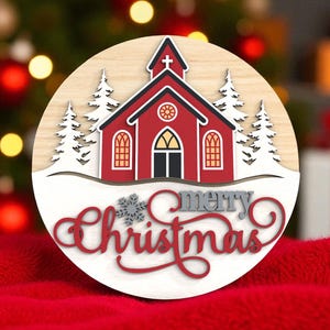 Puede incluir: Adorno navide&ntilde;o redondo de madera con una iglesia roja con detalles blancos, nieve y &aacute;rboles de hoja perenne. Las palabras "merry Christmas" est&aacute;n escritas en una fuente decorativa. El fondo est&aacute; borroso, con una superficie roja.
