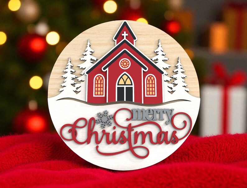 Puede incluir: Adorno navide&ntilde;o redondo de madera con una iglesia roja con detalles blancos, nieve y &aacute;rboles de hoja perenne. Las palabras "merry Christmas" est&aacute;n escritas en una fuente decorativa. El fondo est&aacute; borroso, con una superficie roja.