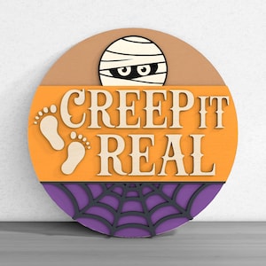 Op de afbeelding: Een rond houten Halloween-bord met een mummie die over de bovenkant kijkt. Het bord toont de tekst "CREEP IT REAL" in een speels lettertype, met een spinnenwebontwerp aan de onderkant en voetafdrukken aan de linkerkant. Het bord is oranje, paars, bruin en wit.