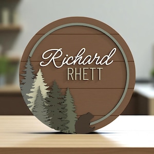 Peut inclure: Un panneau rond en bois avec le nom "Richard" en écriture cursive et "RHETT" en lettres majuscules. Le panneau présente une scène de forêt avec des arbres et une silhouette d'ours, le tout dans des tons de gris et de marron.