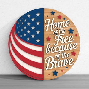 Puede incluir: Un letrero redondo de madera con el diseño de la bandera estadounidense a la izquierda y el texto "Home of the Free because of the Brave" a la derecha. El letrero es rojo, blanco y azul con acentos de estrellas.