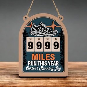 Op de afbeelding: Houten hardlooplogbord met een zwart-witte hardloopschoenafbeelding en de tekst "9999 MILES RUN THIS YEAR". Het bord heeft een teal achtergrond en een houten frame, met een touwtje om op te hangen.