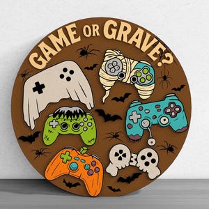 May include: Round wooden sign with the text "GAME or GRAVE?" featuring Halloween-themed game controllers. The controllers are designed as a ghost, mummy, Frankenstein, and pumpkin. Spiders and bats are also included.