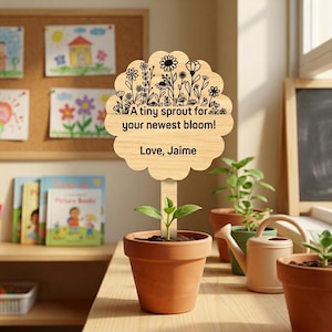 Op de afbeelding: Een houten plantenlabel met een bloemenontwerp en de tekst "A tiny sprout for your newest bloom! Love, Jaime." staat in een terracotta pot met een kleine plant. Andere potten en een gieter staan op de achtergrond.