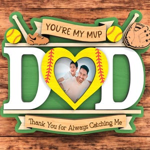 Op de afbeelding: Een houten fotolijst met honkbalthema met het woord "DAD" in grote witte letters. De lijst bevat honkballen, een handschoen en de tekst "You're my MVP" en "Thank You for Always Catching Me". Een foto van een vader en kind staat in de hartvormige lijst.