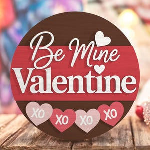 Könnte beinhalten: Rundes Holzschild mit dem Schriftzug "Be Mine Valentine" in weißer Schrift. Das Schild hat einen braunen oberen Teil und einen roten Streifen in der Mitte. Vier rosa Herzen mit "XO" befinden sich am unteren Rand. Das Schild liegt auf einer Holzoberfläche.