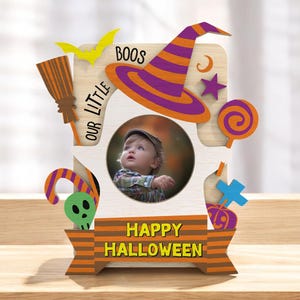 以下が含まれることがあります： ハロウィーンをテーマにした木製フォトフレーム。写真用の円形の開口部があります。フレームは魔女の帽子、ほうき、ロリポップ、そして「BOOS」、「OUR LITTLE」、「HAPPY HALLOWEEN」の文字で飾られています。フレームはオレンジ、紫、黄色で塗装されています。