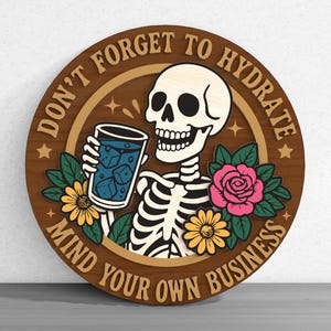 May include: Round wooden sign featuring a skeleton holding a drink with ice cubes, surrounded by flowers. The sign has the text "DON'T FORGET TO HYDRATE" and "MIND YOUR OWN BUSINESS" in a circular design.