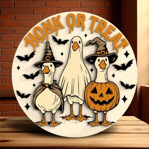Puede incluir: Decoraci&oacute;n redonda de Halloween de madera con tres gansos de dibujos animados. Uno est&aacute; disfrazado de fantasma, otro de bruja y el tercero de calabaza. Las palabras "HONK OR TREAT" est&aacute;n en la parte superior, con murci&eacute;lagos y estrellas.