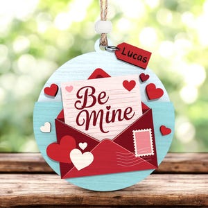 Könnte beinhalten: Valentinstags-Ornament mit hellblauem kreisförmigem Hintergrund, rotem Umschlag und Herzen. Der Umschlag zeigt eine Notiz mit der Aufschrift "Be Mine". Ein kleines rotes Etikett mit dem Namen "Lucas" ist angebracht.