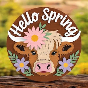 May include: A circular wooden sign featuring a cow's face and the text "Hello Spring". The cow is adorned with pink and purple flowers and green leaves. The sign has a brown background. The text is in white.