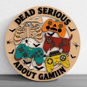 May include: A round wooden sign with the words "DEAD SERIOUS ABOUT GAMIN" in black. The sign features a skeleton, a pumpkin, and three game controllers in teal, red, and yellow, with bats and spiders.