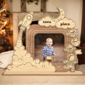 Puede incluir: Un marco de fotos de madera con forma de cohete con un diseño de nube y luna. El marco tiene espacios para escribir el nombre, el lugar, el peso, la altura y la hora de nacimiento del bebé.